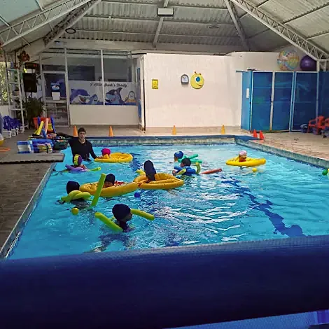 Varios niños con flotadores practican en una piscina cubierta, acompañados por una instructora dentro del agua.